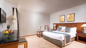 A hotel room with a bed, bedside tables, a desk with fruit, a TV, and artwork on the wall.