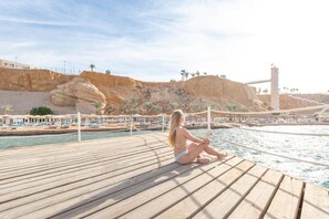 Eine Holzterrasse mit einer Person am Wasser, einer felsigen Klippe und einer Brücke im Hintergrund.