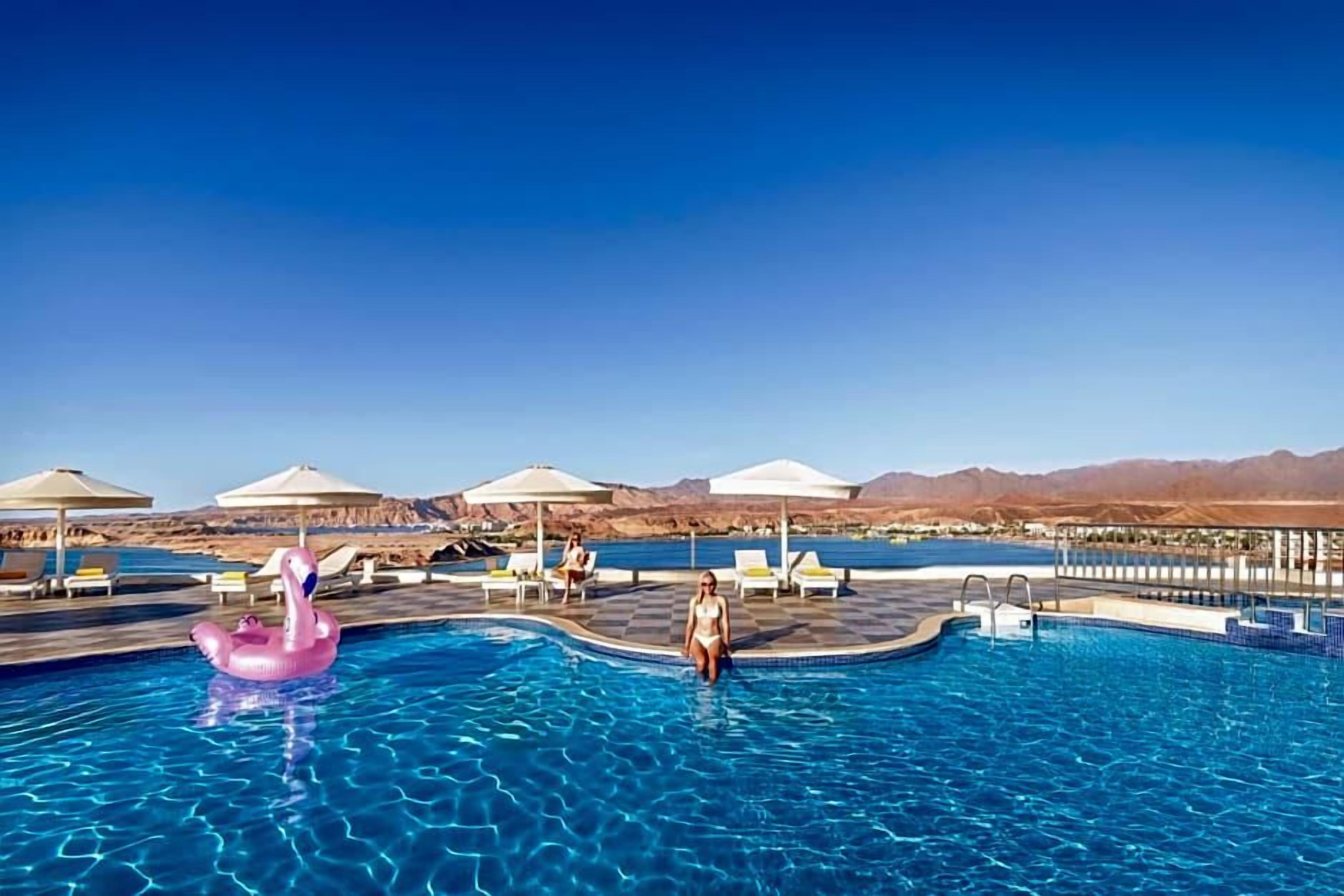 En swimmingpool med liggestole og parasoller, omgivet af bjerge.