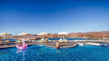 En swimmingpool med liggestole og parasoller, omgivet af bjerge.