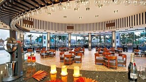 A spacious indoor dining area with multiple tables and chairs, large windows offering views of palm trees, and a ceiling with hanging light fixtures. - Albatros Sharm Resort By Pickalbatros (Sharm El Sheikh)