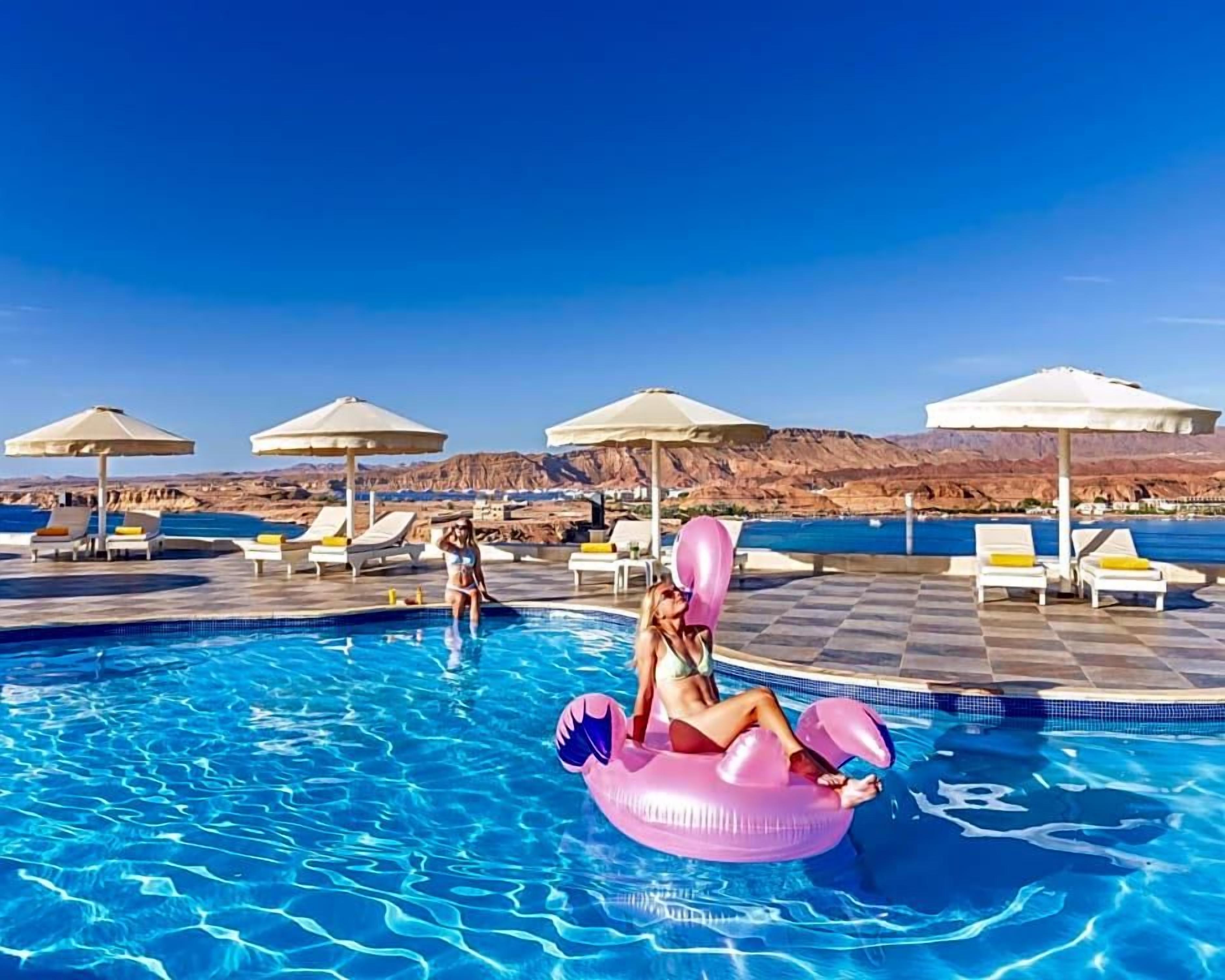 A pool area with lounge chairs and umbrellas, a pool, and a mountainous backdrop.