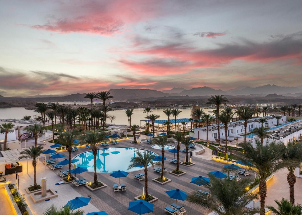 A resort with a central pool surrounded by palm trees and lounge chairs, overlooking a body of water and mountains.