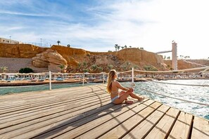 Une terrasse en bois où est assise une personne, donnant sur un espace piscine avec des chaises longues et des parasols, et avec une falaise rocheuse en arrière-plan.