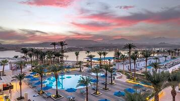 A resort with a large pool surrounded by palm trees and lounge chairs, set against a backdrop of mountains and a sunset sky.
