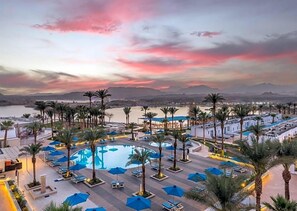Un complexe hôtelier doté d’une grande piscine entourée de palmiers et de chaises longues, le tout situé dans un cadre magnifique avec des montagnes en arrière-plan et un ciel au coucher du soleil.