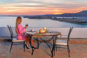 Une femme est assise à une table ronde, d’où l’on peut admirer un lac et des montagnes au coucher du soleil.
