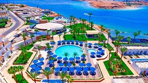 Ein Resort mit einem zentralen Swimmingpool, umgeben von Liegestühlen und Palmen, eine Straße, die sich durch das Anwesen schlängelt, und ein Strand im Hintergrund.