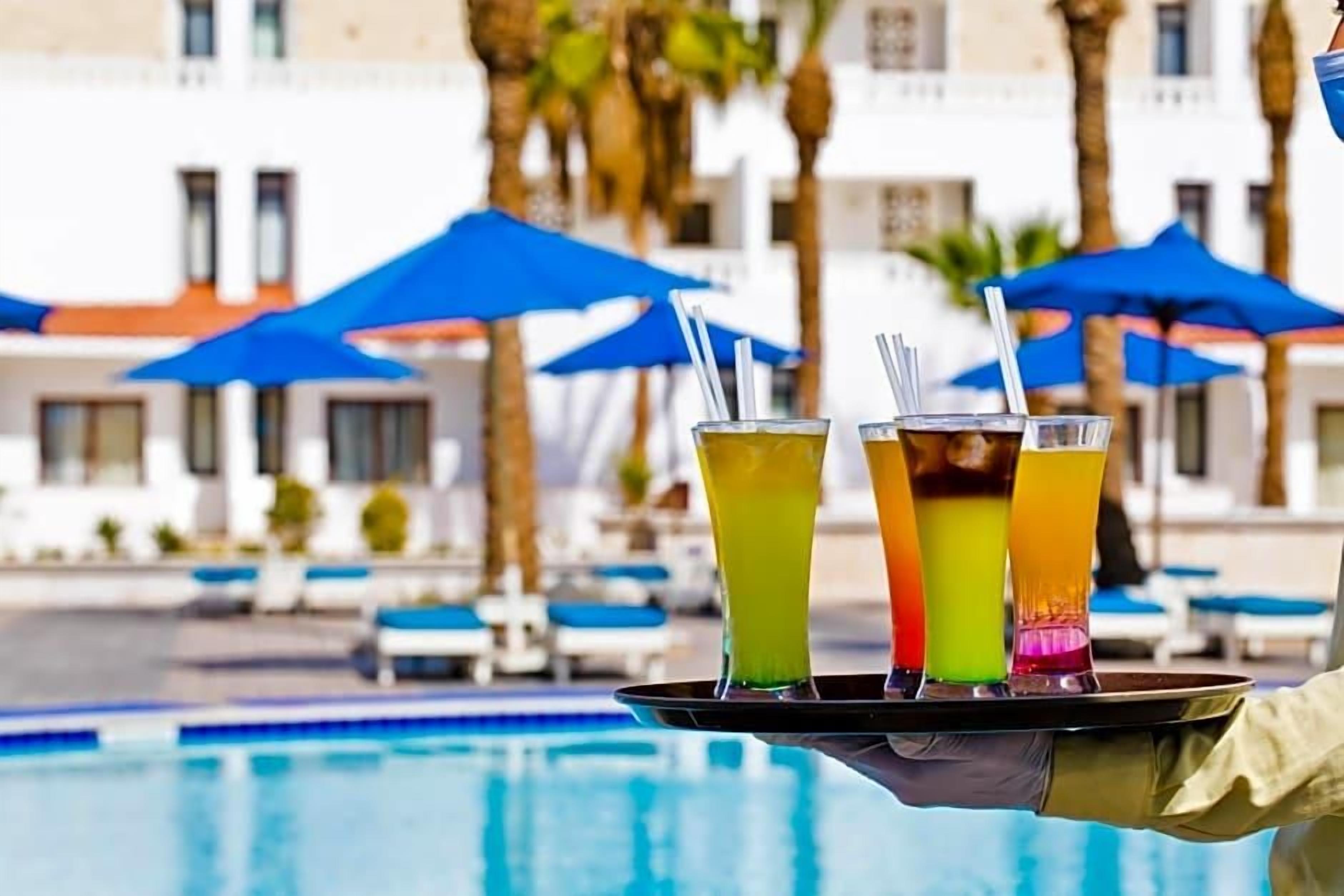 A tray with three colorful drinks, a pool, and a building with blue umbrellas in the background.