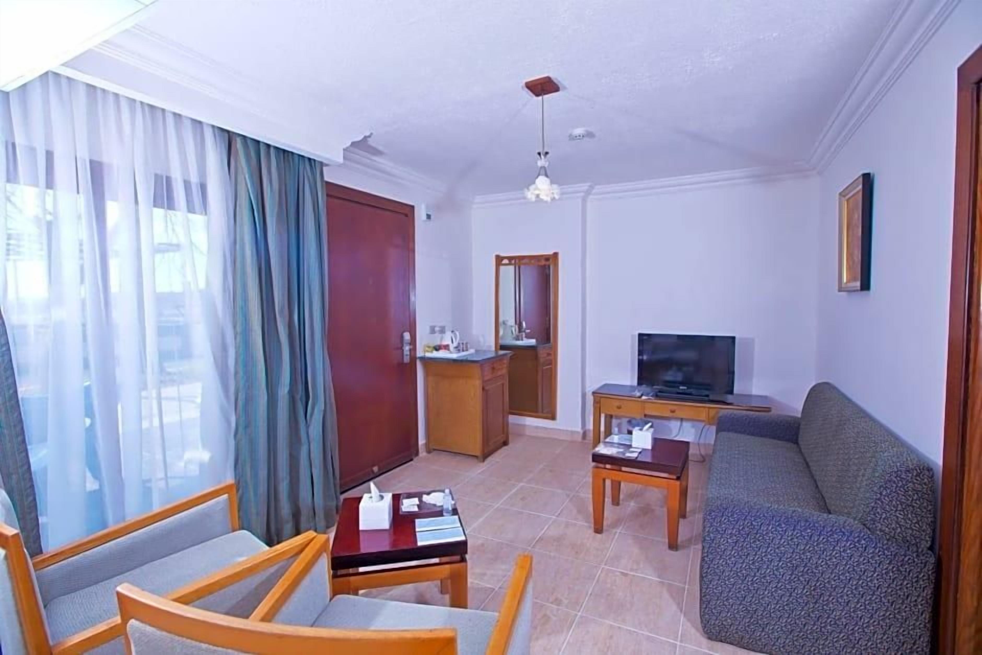 A living room with a sofa, chairs, a TV, a small table, and a kitchen area with a mirror and cabinets.