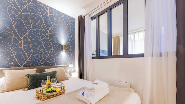 A hotel room with a bed, a wall with a tree branch pattern, a window with curtains, and a bedside table with a bottle and glasses. - Beautiful Apartment at Gare de L'est Republique (Paris)