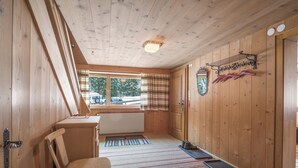 Interior de una cabaña de madera con una silla, un radiador, una ventana con cortinas y un perchero con sombreros.