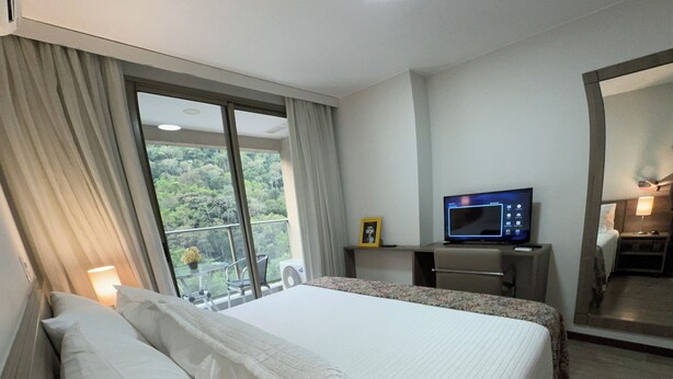 A hotel room with a large bed, a desk with a computer, a TV, and a balcony with a view of greenery. - Comfort & king bed near Riocentro and Olympic Park (Jacarepaguá Rio de Janeiro)