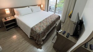 A hotel room with a large bed, a desk with a chair, a TV, and a balcony with a view of buildings and trees. - Comfort & king bed near Riocentro and Olympic Park (Jacarepaguá Rio de Janeiro)