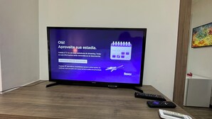 A television on a wooden table displaying a screen with text in Portuguese, accompanied by a remote control and a mobile device. - Comfort & king bed near Riocentro and Olympic Park (Jacarepaguá Rio de Janeiro)