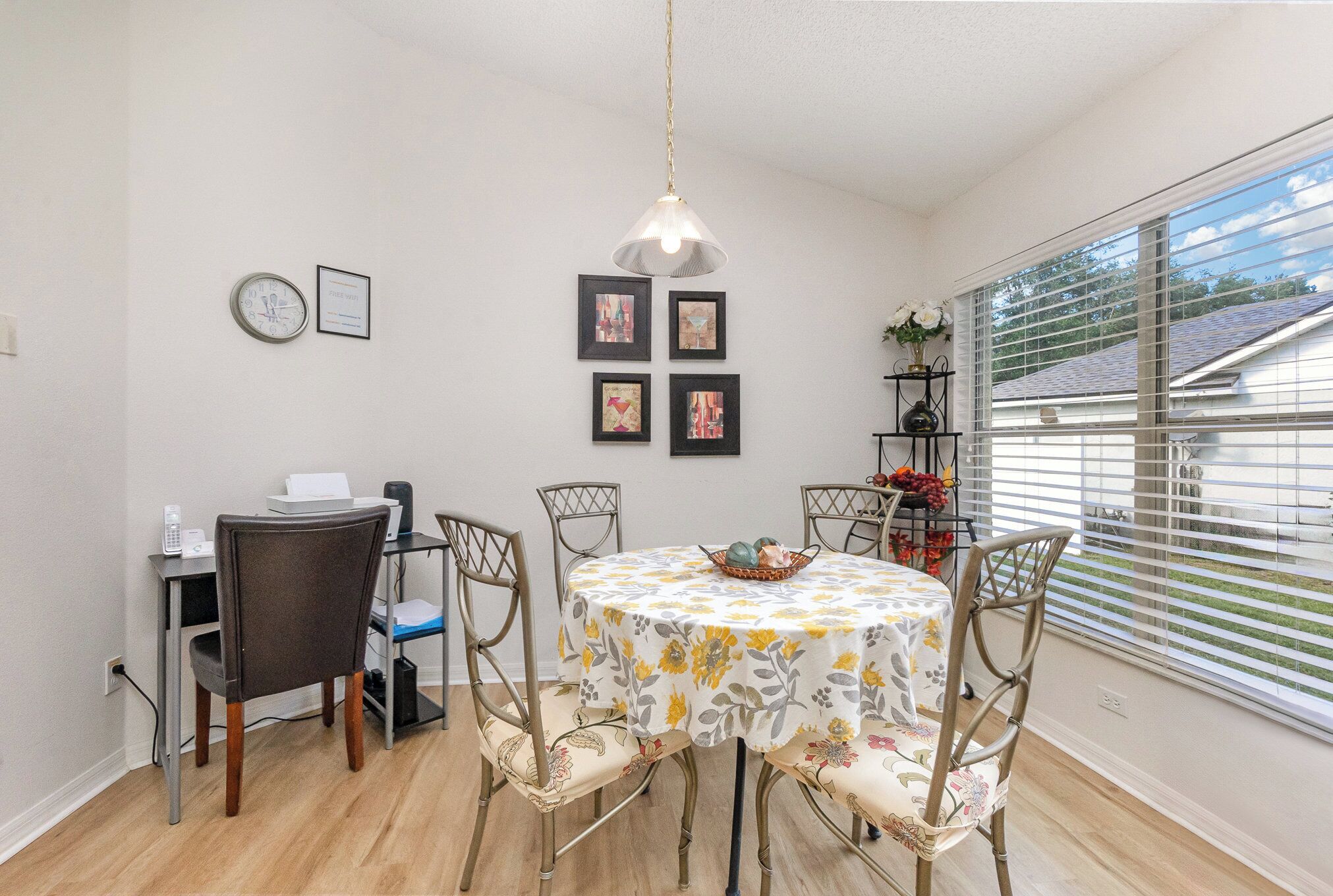 A dining area with a round table, chairs, a clock, framed pictures, and a window with blinds.