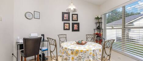 A dining area with a round table, chairs, a clock, framed pictures, and a window with blinds.
