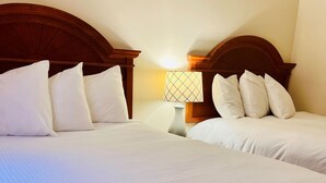 A hotel room with two beds, each with white linens and multiple pillows. There is a nightstand with a lamp between the beds.
