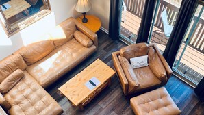 A spacious living area with a brown leather sofa set, a wooden coffee table, and a lamp. A wooden staircase is visible in the foreground.