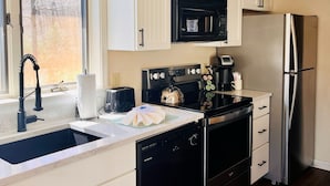 A kitchen with white cabinets, a black microwave, a stainless steel refrigerator, a dishwasher, and a window with a view of trees.