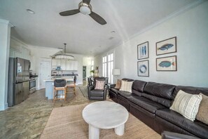 Uma cozinha moderna com eletrodomésticos de aço inoxidável, uma sala de jantar com mesa e cadeiras, uma sala de estar com sofá de couro e mesinha de centro, e um ventilador de teto.
