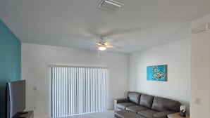 Ein Wohnzimmer mit einem grauen Ecksofa, einem Flachbildfernseher auf einem Holzständer, einem Deckenventilator und einem Gemälde an der Wand.