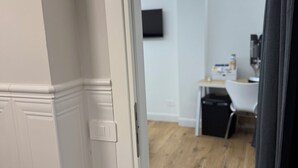 A room with a wooden floor, white walls, a flat-screen TV mounted on the wall, a small table with a chair, and a black trash bin.