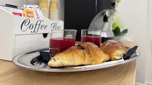 A breakfast tray with croissants and two glasses of red juice, a coffee bar, and stacked cups and condiments.