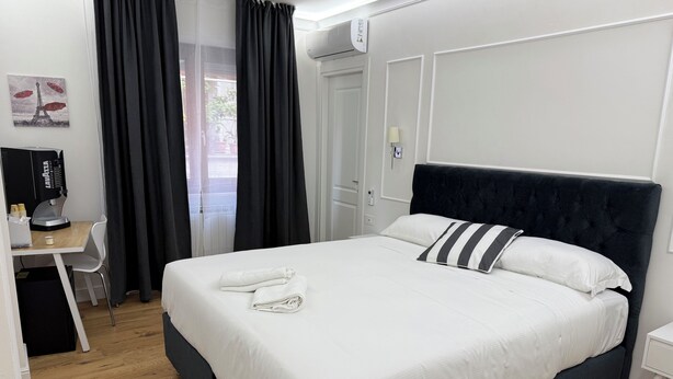 A neatly made bed with white linens, a dark headboard, and striped pillows. There is a desk with a computer and a coffee machine, and a window with black curtains.