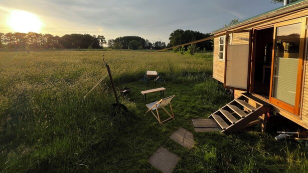 A small wooden cabin with a sliding door, a small table, and a chair outside, set in a grassy field with a clear sky. - Beautiful circus wagon in a secluded location  (Oetzen)