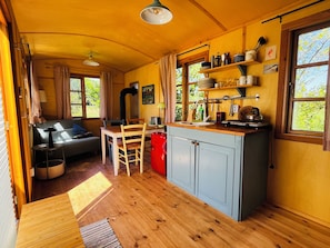 A cozy kitchen with wooden cabinets, a dining table, and chairs. There is a window with curtains, a sink, and various kitchen utensils.