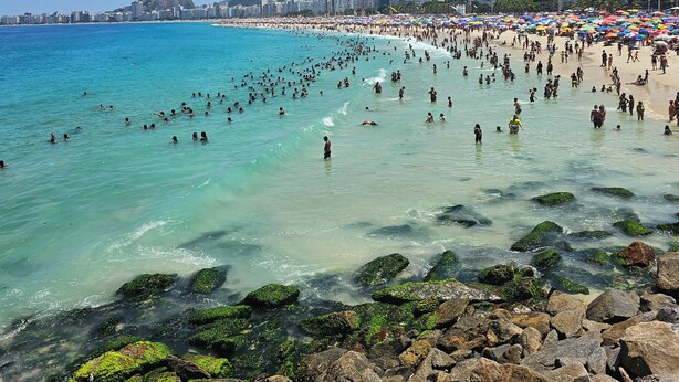 A crowded beach with clear turquoise water, rocky shoreline, and a cityscape in the background.