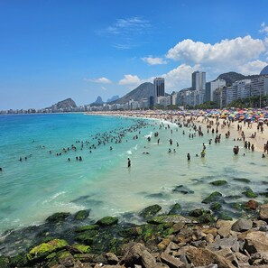 A crowded beach with clear turquoise water, rocky shoreline, and a cityscape in the background.