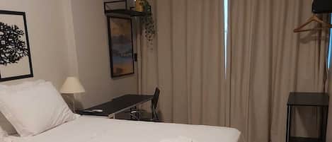 A hotel room with a large bed, a desk, a chair, a lamp, and a view of the outdoors through curtains.
