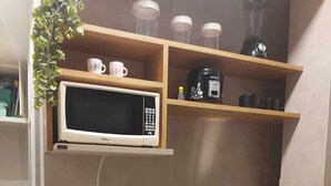 A compact kitchen with a built-in microwave, a small refrigerator, a sink with a dish rack, and wooden shelves above the counter.