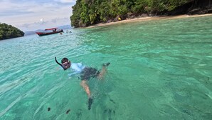 Una persona practica snorkel en aguas turquesas cristalinas, con un barco y una isla boscosa al fondo.