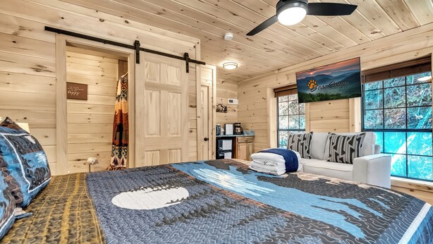 A cozy cabin interior with a bed, a sofa, a ceiling fan, and a large window offering a view of the outdoors. - Renovated Barn with Creek and Waterfall (Cosby)