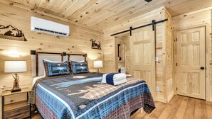 A wooden-paneled bedroom with a bed, two bedside lamps, a wall-mounted air conditioner, and a door with a sliding mechanism. - Renovated Barn with Creek and Waterfall (Cosby)