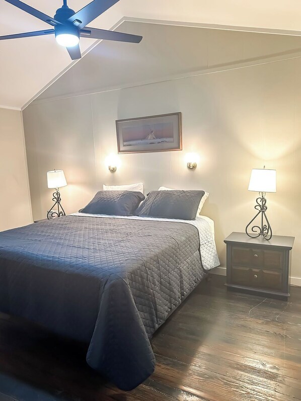 A bedroom with a bed, two bedside tables with lamps, a ceiling fan, and a framed picture on the wall.