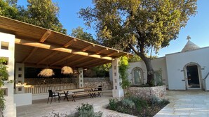 Un espace extérieur aménagé, abrité et doté de bancs et de guirlandes lumineuses, entouré de murs en pierre et d’un jardin où poussent différentes espèces de plantes.