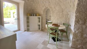 Une pièce aux murs de pierre, avec un meuble blanc, une table entourée de chaises et une porte donnant sur l’extérieur.