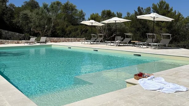 Une piscine d’un bleu éclatant, bordée de chaises longues et d’ombrelles, entourée d’arbres et d’un mur de pierre.