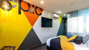 A modern bedroom with a geometric wall design, a flat-screen TV, an air conditioning unit, and a bed with yellow and gray bedding. - Epic Boutique Hotel (Medellín)
