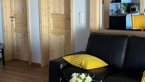 Een moderne woonkamer met een glazen salontafel, een zwarte bank, houten deuren en een keuken op de achtergrond.