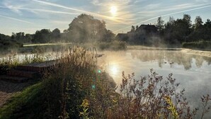 Een rustig stadsgezicht aan een meer met een houten steiger, kalm water dat de lucht spiegelt en omliggende begroeiing.