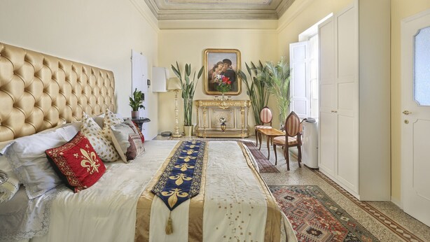 Une chambre à coucher avec une tête de lit capitonnée, un lit recouvert d’une literie à motifs, une cheminée, un tableau au-dessus, et une vue sur l’extérieur par une porte.
