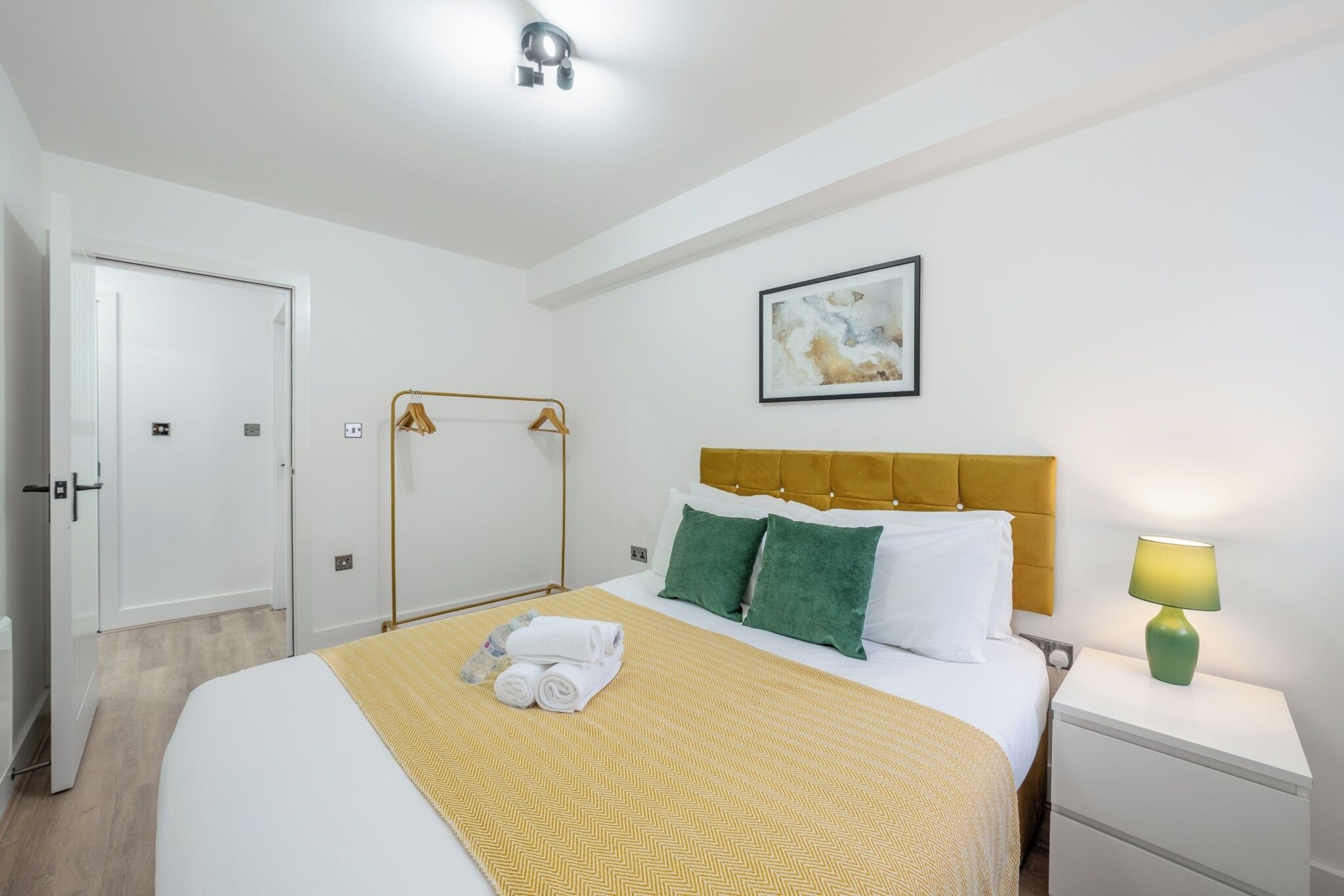 A hotel room with a bed, a nightstand, a wall-mounted lamp, a painting, and a door.