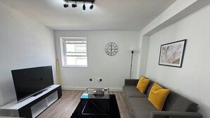 A modern living room with a flat-screen TV, a grey sofa with yellow cushions, a black coffee table, and a large window providing natural light. - Deluxe 3 Bedstratford Apartment Olympicparko2excel (London)