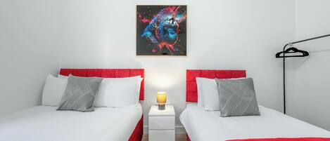 Une chambre d’hôtel avec deux lits, chacun doté d’une tête de lit rouge et de draps blancs. Il y a une table de chevet avec une lampe entre les deux lits, et un tableau abstrait encadré est accroché au mur au-dessus de la table de chevet.