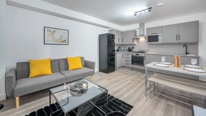 A modern apartment with a grey sofa, a dining table with chairs, a kitchen area with grey cabinets and appliances, and a black-framed picture on the wall. - Deluxe 3 Bedstratford Apartment Olympicparko2excel (London)